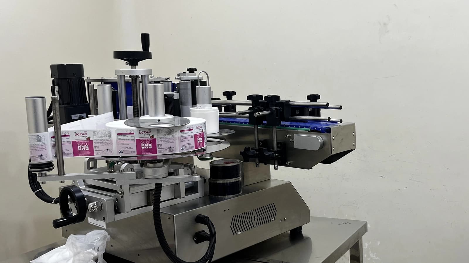 Stainless-steel labelling machine with a conveyor and a roll of printed UCRAN Syrup labels loaded on its reel