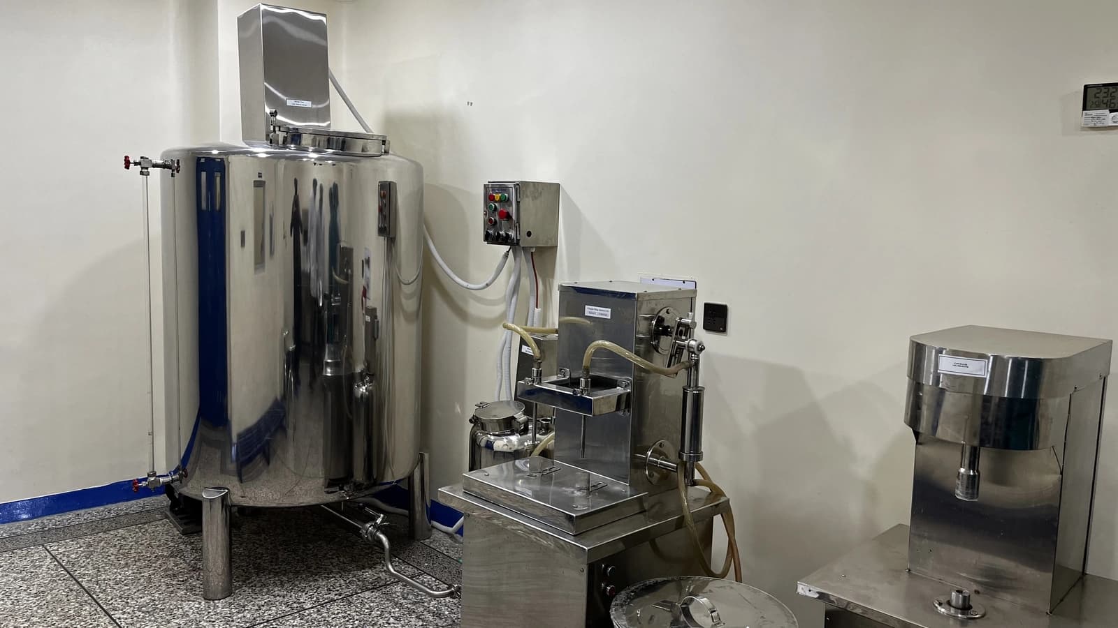 Large stainless-steel tank with a sight-glass level gauge and control box, next to bench-top stainless-steel liquid filling and cap-sealing machines