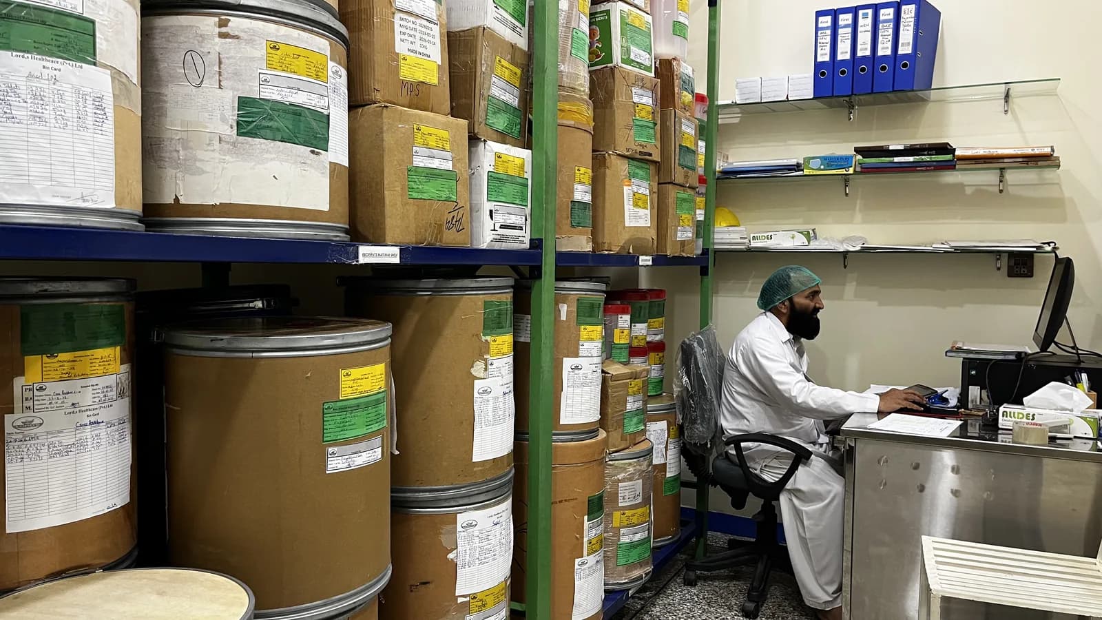 Raw-material store with labelled fibre drums and cartons on blue and green racking, and a staff member in a hairnet working at the store desk